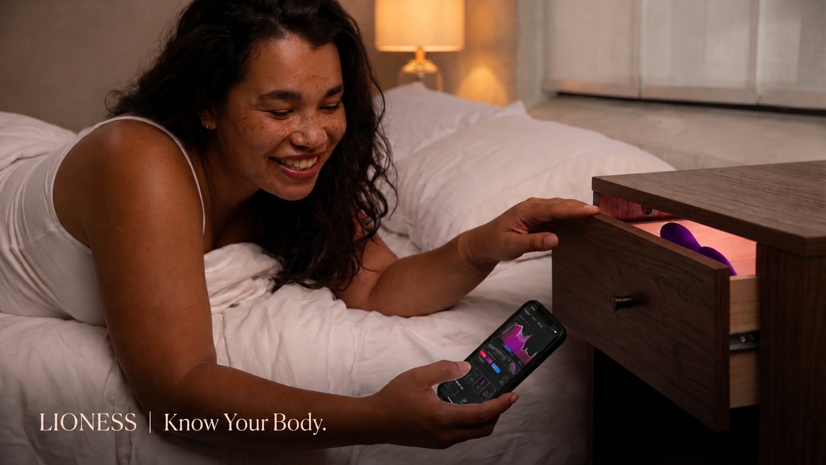 Woman in bed smiling at the Lioness app on her phone, next to her nightstand